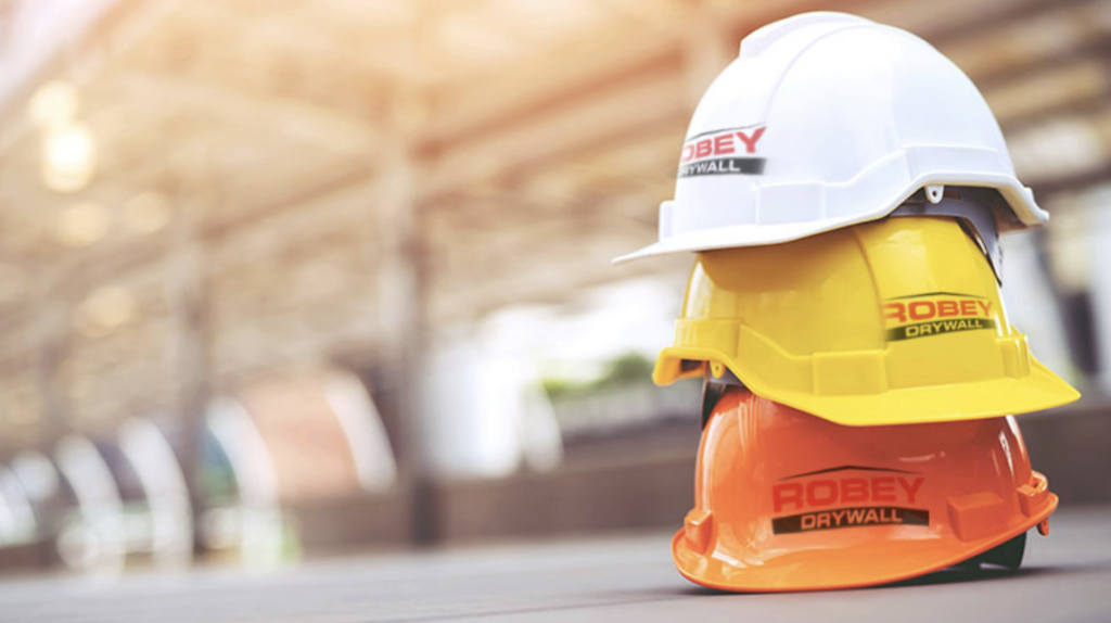 stack of white, yellow, and orange OSHA safety helmets branded with Robey Drywall's logo